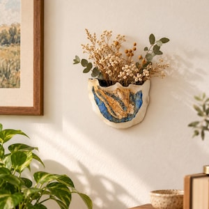 May include: A wall-mounted ceramic planter with an irregular, wavy rim. The exterior features a white base with a textured, abstract design in shades of deep blue and golden brown. It is filled with a bouquet of dried flowers and green eucalyptus.