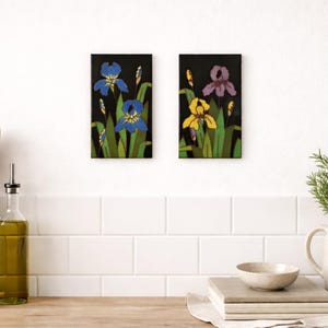 May include: Two framed floral art pieces. One features blue irises, the other yellow and purple irises, all against a black background. Green leaves surround the flowers. The art is displayed on a white wall above a wooden countertop.