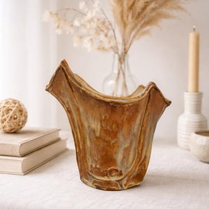 May include: A handcrafted, brown ceramic vase with a unique, flared shape. The vase has a textured surface with a mix of brown and beige tones. Decorative elements include a small, round ball and a candle.