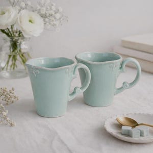 May include: Two light blue ceramic mugs with ornate handles and rims. The mugs are on a white surface, with a small plate, gold spoon, and sugar cubes nearby. A vase of white flowers is in the background.
