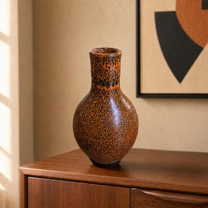 May include: A ceramic vase with a bulbous body and a narrow neck, featuring a speckled pattern of black dots over a vibrant orange background. The surface has a glossy finish, and the vessel stands upright with a rounded base and a flared rim.