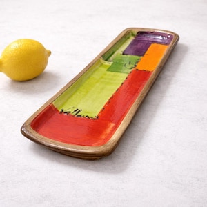 May include: A rectangular ceramic serving tray with a colorful abstract design. The tray features a wooden-look border and a vibrant interior with red, green, orange, and purple blocks. The word "Vallauris" is written in black script. A yellow lemon is in the background.