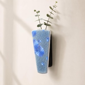May include: A wall-mounted ceramic vase with a triangular shape and a light blue crystalline glaze featuring dark blue flower-like patterns and a fine crackle texture. The vase holds a single green eucalyptus stem with small round leaves.