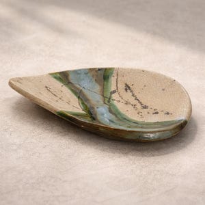 Op de afbeelding: Een druppelvormige keramische schaal met een beige basis en een gekraakt oppervlak. De schaal heeft blauwe en groene strepen, met donkerbruine accenten. De randen zijn iets verhoogd en het algehele ontwerp is organisch en artistiek.