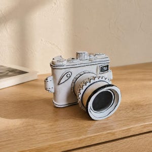 May include: A white ceramic camera sculpture featuring hand-painted black outlines that create a two-dimensional, cartoon-like appearance. The object includes a prominent lens, a top dial, and a rectangular viewfinder, all rendered in a flat, graphic style.