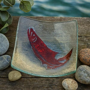 May include: Square glass dish with a red fish design. The dish has a slightly curved shape and a clear, textured surface. The fish is a vibrant red color, and the dish has a light blue and beige background. The edges of the dish are a light green color.