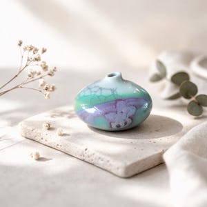 May include: A small, ceramic vase with a rounded shape and a narrow opening. The vase features a gradient of colors, including light blue, green, and purple. The surface has a crackle glaze effect. The vase is displayed on a rectangular stone.