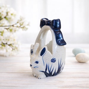 May include: A ceramic Easter basket shaped like a bunny, with a navy blue bow on the handle. The basket is white with blue accents, including the bunny's ears and floral designs. The basket is sitting on a white wooden surface.