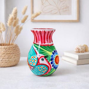 Könnte beinhalten: Eine handbemalte Keramikvase mit einem lebendigen Design. Die Vase hat einen roten und rosa Rand, einen türkisfarbenen Körper mit einem Vogel- und Blumenmuster und grüne Blätter. Die Vase ist ein dekorativer Artikel.