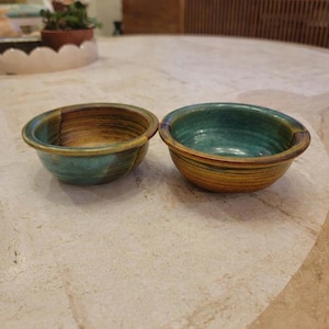 Puede incluir: Dos cuencos de cerámica hechos a mano con una estética rústica. Los cuencos son redondos con un perfil bajo y presentan una mezcla de tonos turquesa, marrón y amarillo. Los cuencos probablemente se utilizan para servir alimentos o como piezas decorativas.