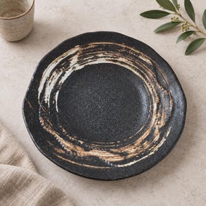 May include: A round ceramic plate with a dark black, speckled base featuring a prominent, circular brushstroke pattern in shades of tan and white. The plate has an irregular, organic edge and a textured surface, showcasing a hand-crafted appearance.