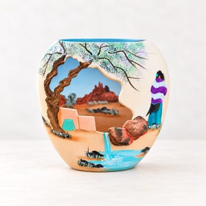 May include: A rounded ceramic vase featuring a hand-painted desert landscape scene. The design includes a cutout section revealing a painted view of red rock formations, a gnarled tree, a small structure, and a figure wearing a striped garment.
