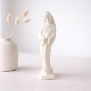 May include: A cream-colored ceramic statue of a woman in a flowing gown, with her hands clasped in front of her. The statue is on a square base. A small white vase with dried flowers is in the background.