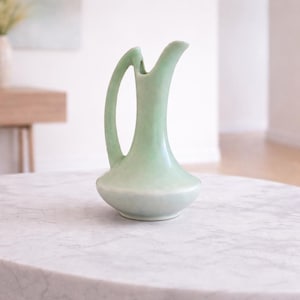 May include: A pale green ceramic jug with a curved handle and a spout. The jug has a bulbous base and a slender neck. It is sitting on a marble surface. The jug is likely used for serving beverages.