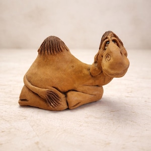 May include: A small, handcrafted ceramic camel figurine in a seated position. The camel is a warm, earthy brown color with detailed carving on the hump, head, and tail. The figurine has a whimsical, expressive face.