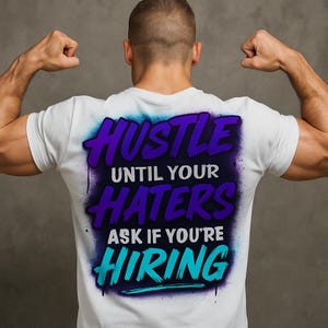 May include: White t-shirt with the text "HUSTLE UNTIL YOUR HATERS ASK IF YOU'RE HIRING" in purple and teal. The text is in a graffiti-style font with a blue and purple spray paint effect. The person is flexing their arms.