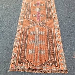 May include: A long, narrow runner rug featuring a vibrant orange base with geometric patterns in brown, cream, and pink. The design includes repeating central motifs and a triangular border, finished with short, frayed fringe at the bottom edge.