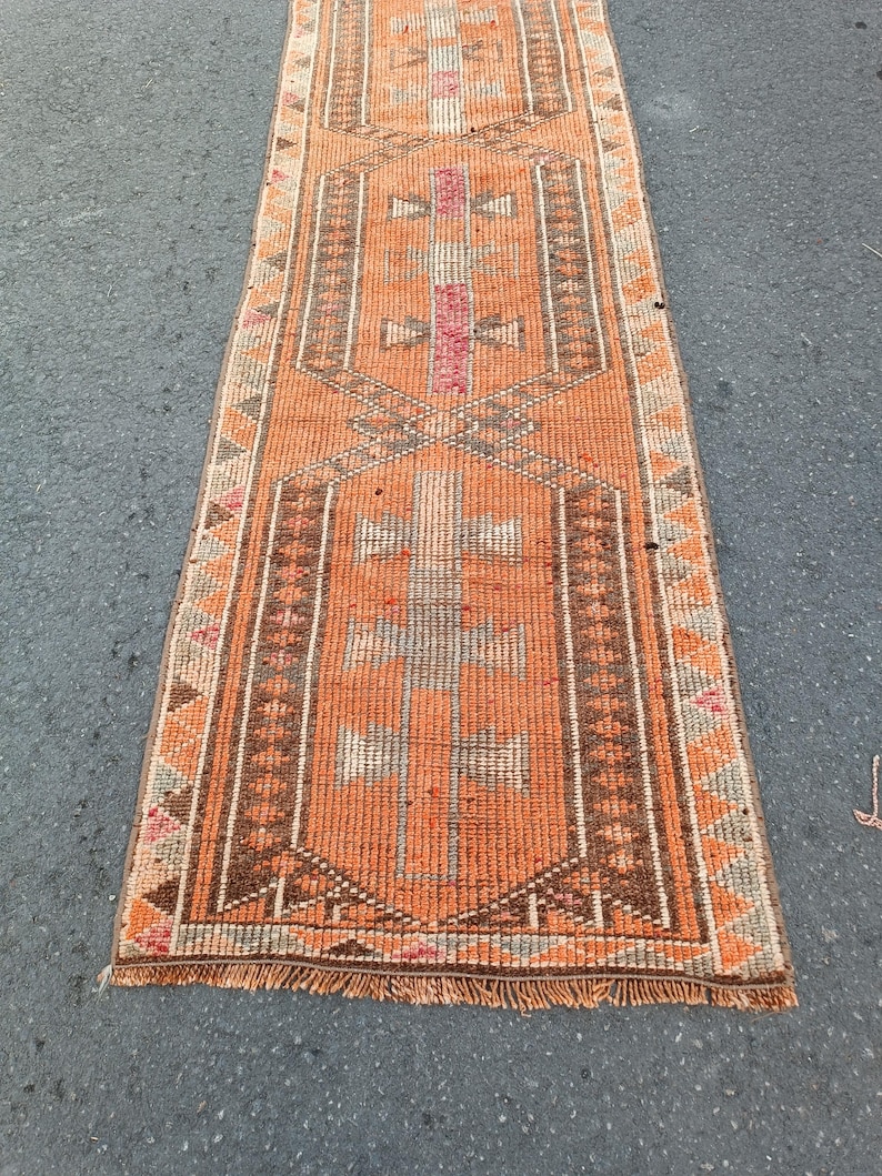 May include: A long, narrow runner rug featuring a vibrant orange base with geometric patterns in brown, cream, and pink. The design includes repeating central motifs and a triangular border, finished with short, frayed fringe at the bottom edge.