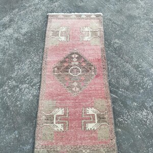 Puede incluir: Una alfombra rectangular de estilo vintage con un campo rosa descolorido. La alfombra presenta patrones geométricos en marrón, beige y blanco. El diseño incluye una forma de diamante central y motivos de esquina estilizados. La alfombra tiene un borde con flecos.