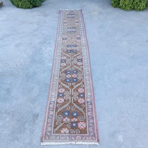 Könnte beinhalten: Ein langer, schmaler Läufer mit einem braunen Mittelfeld und einem sich wiederholenden geometrischen Muster aus blauen und rosafarbenen Blumenmotiven. Der Teppich ist von einer dekorativen Bordüre in Rosa, Grau und Weiß umrahmt und hat weiße Fransen.