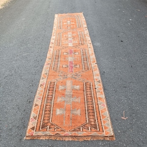 May include: A long, rectangular runner rug featuring a vibrant orange base color with intricate geometric patterns in shades of brown, white, and pink. The rug has a woven texture, a repeating central motif, and short fringe along both ends.