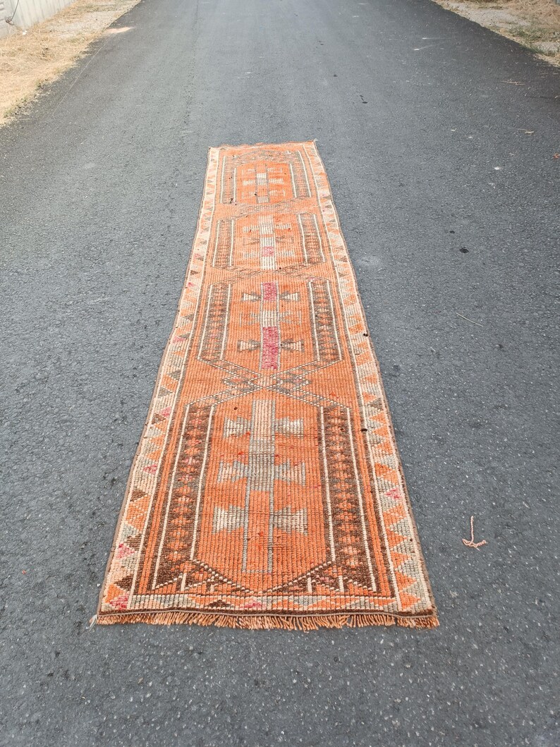 May include: A long, rectangular runner rug featuring a vibrant orange base color with intricate geometric patterns in shades of brown, white, and pink. The rug has a woven texture, a repeating central motif, and short fringe along both ends.