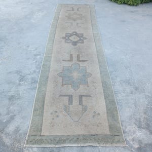 May include: A long, rectangular runner rug featuring a faded beige base with a series of geometric star-like motifs in muted blue and grey tones. The rug has a wide, sage green border and short, light-coloured fringe at both ends of the piece.