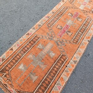 May include: A long, rectangular runner rug featuring a vibrant orange base color with intricate geometric patterns in shades of brown, cream, and muted pink. The design includes repeating diamond-shaped motifs and a fringed edge at the bottom.