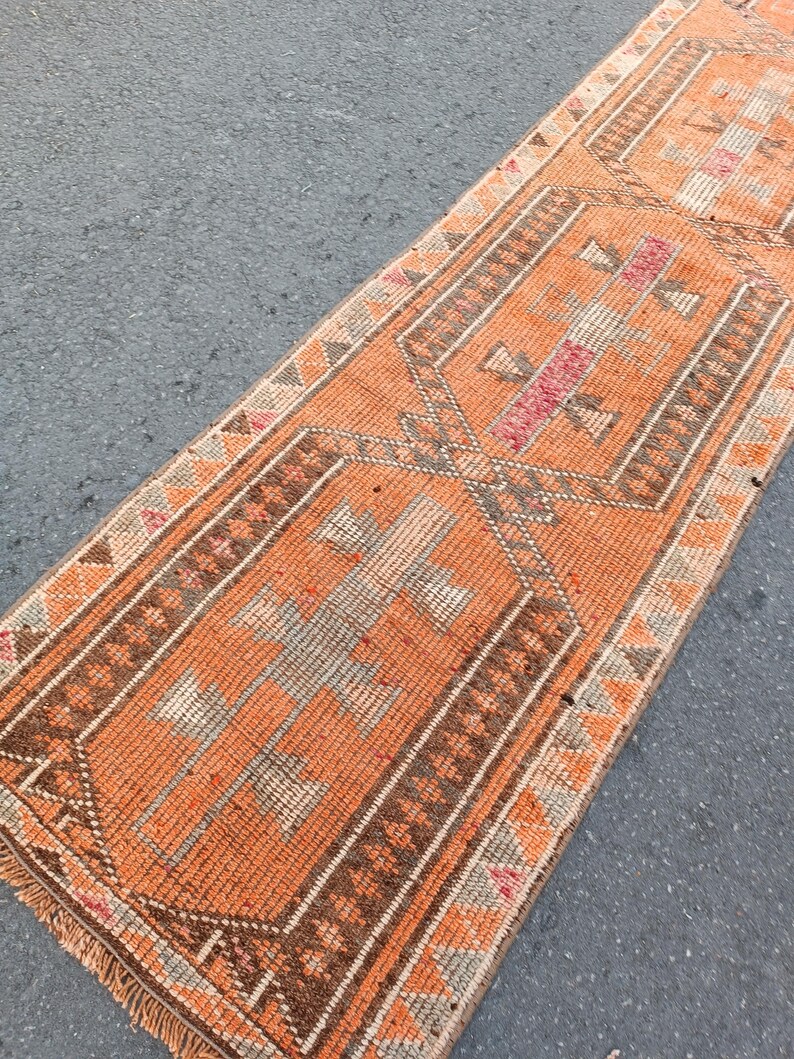May include: A long, rectangular runner rug featuring a vibrant orange base color with intricate geometric patterns in shades of brown, cream, and muted pink. The design includes repeating diamond-shaped motifs and a fringed edge at the bottom.
