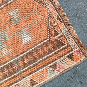 May include: A woven rug featuring a geometric pattern in shades of orange, brown, and cream. The textile has a textured pile surface with a decorative border and a fringed edge, showcasing intricate knotted details throughout the rectangular design.