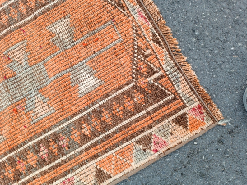 May include: A woven rug featuring a geometric pattern in shades of orange, brown, and cream. The textile has a textured pile surface with a decorative border and a fringed edge, showcasing intricate knotted details throughout the rectangular design.