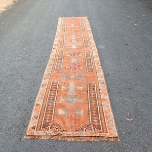 May include: A long, rectangular runner rug featuring a vibrant orange base color with intricate geometric patterns in shades of brown, white, and muted pink. The rug has a repeating central motif and a fringed edge along the short ends.