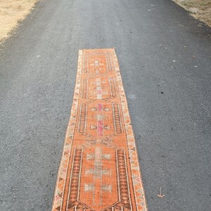 May include: A long, rectangular runner rug featuring a vibrant orange base color with intricate geometric patterns in shades of brown, cream, and pink. The rug has a symmetrical design with repeating central motifs and a fringed edge at the bottom.