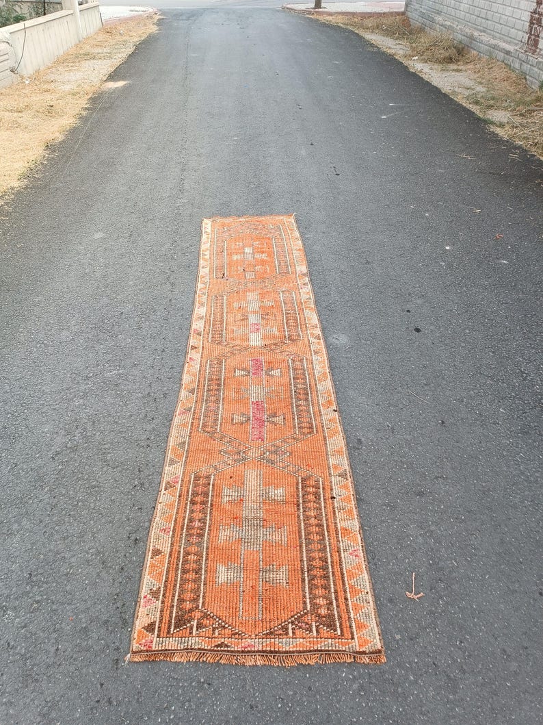 May include: A long, rectangular runner rug featuring a vibrant orange base color with intricate geometric patterns in shades of brown, cream, and pink. The rug has a symmetrical design with repeating central motifs and a fringed edge at the bottom.