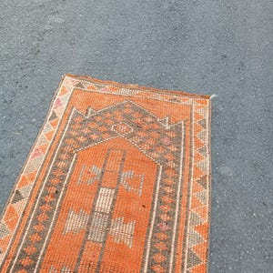 May include: A rectangular woven rug featuring a vibrant orange field with a central dark brown mihrab arch design. The border displays a repeating geometric triangle pattern in brown and orange, with small accents of pink and cream throughout the weave.
