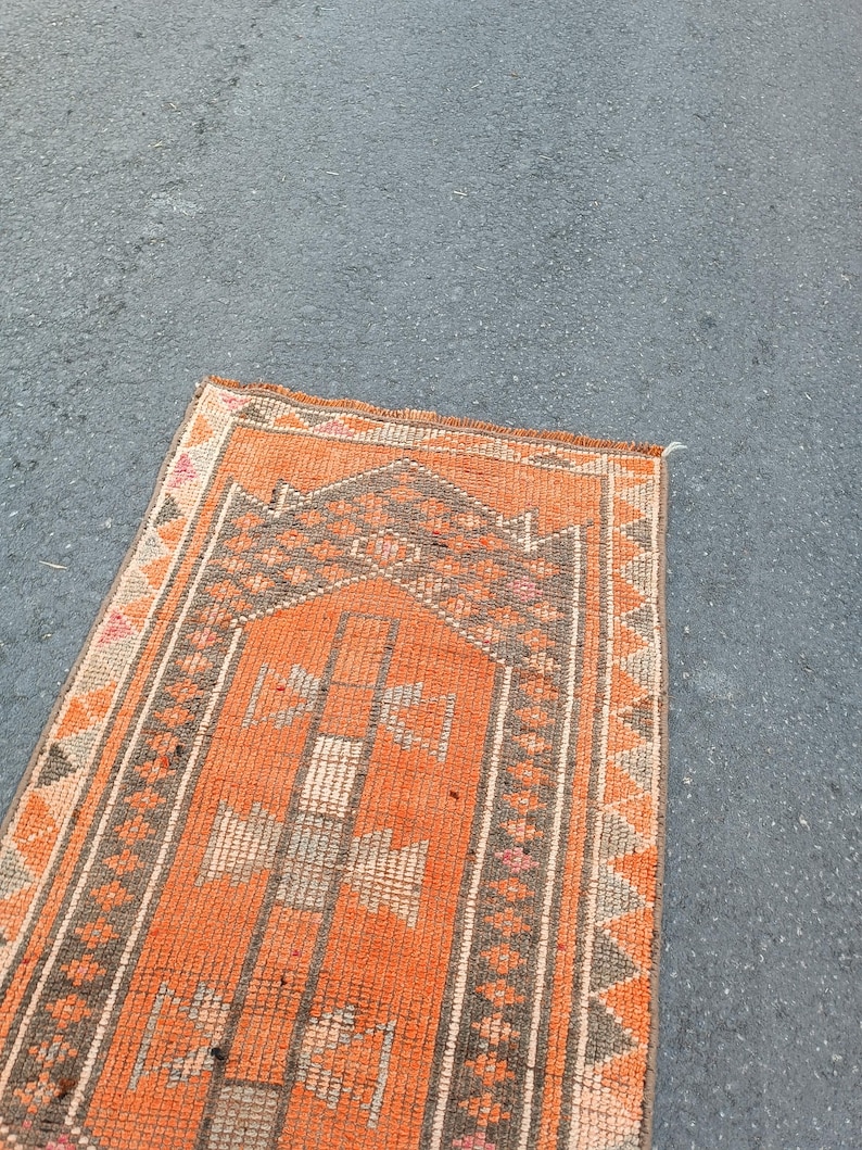 May include: A rectangular woven rug featuring a vibrant orange field with a central dark brown mihrab arch design. The border displays a repeating geometric triangle pattern in brown and orange, with small accents of pink and cream throughout the weave.