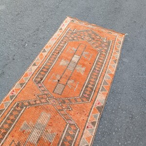 May include: A rectangular runner rug featuring a vibrant orange field with a central geometric pattern in shades of gray, white, and muted pink. The border displays a repeating triangular motif in orange, gray, and white, framing the design.