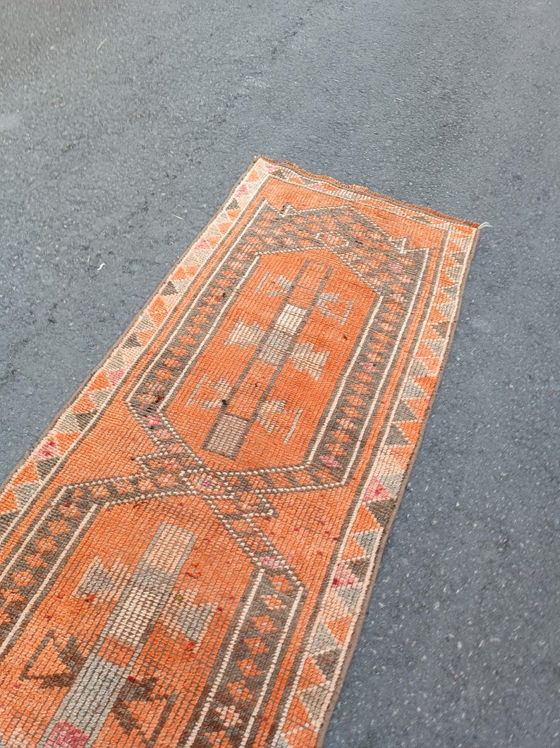 May include: A rectangular runner rug featuring a vibrant orange field with a central geometric pattern in shades of gray, white, and muted pink. The border displays a repeating triangular motif in orange, gray, and white, framing the design.