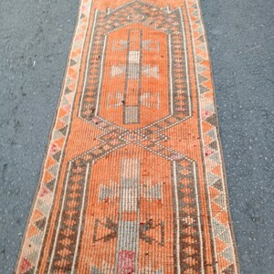 May include: A long, narrow runner rug featuring a vibrant orange background with geometric patterns in brown, gray, and muted pink. The design includes two central hexagonal medallions with stylized cross motifs and a border of repeating triangles.