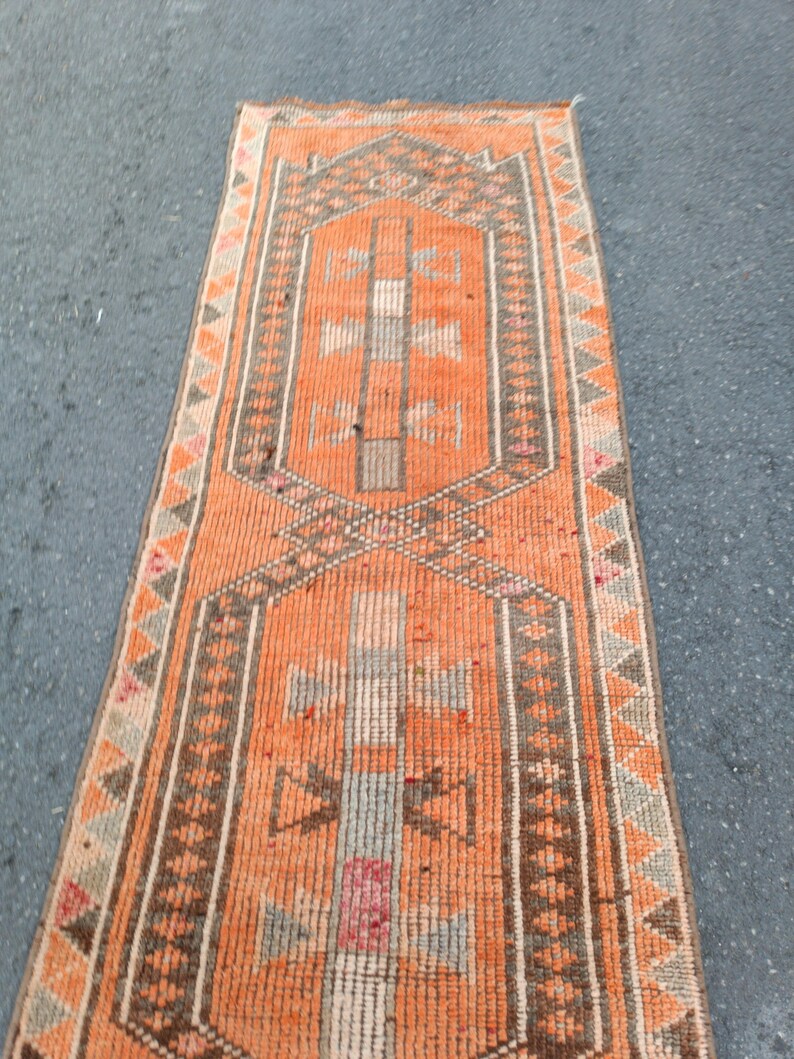 May include: A long, narrow runner rug featuring a vibrant orange background with geometric patterns in brown, gray, and muted pink. The design includes two central hexagonal medallions with stylized cross motifs and a border of repeating triangles.