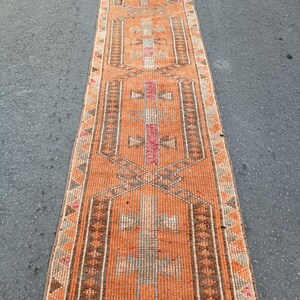 May include: A long, narrow runner rug featuring a vibrant orange base color with repeating geometric patterns in brown, cream, and pink. The design includes central hexagonal motifs and a border decorated with small, repeating triangular shapes.