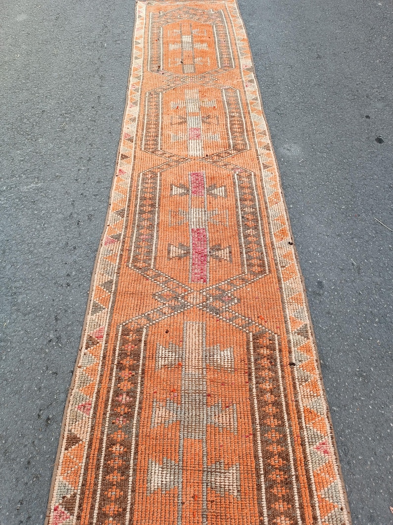 May include: A long, narrow runner rug featuring a vibrant orange base color with repeating geometric patterns in brown, cream, and pink. The design includes central hexagonal motifs and a border decorated with small, repeating triangular shapes.