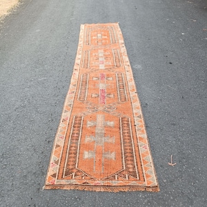 May include: A long, rectangular runner rug featuring a vibrant orange base color with intricate geometric patterns in brown, white, and muted pink. The rug has a repeating central motif, a decorative border with triangular accents, and short fringe.