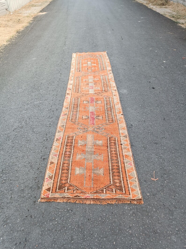 May include: A long, rectangular runner rug featuring a vibrant orange base color with intricate geometric patterns in brown, white, and muted pink. The rug has a repeating central motif, a decorative border with triangular accents, and short fringe.
