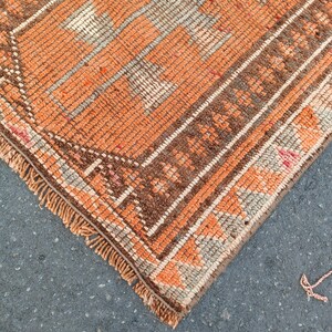 May include: A woven rug featuring a vibrant orange and brown geometric pattern with a central medallion design. The rug has a textured pile, a decorative border with repeating motifs, and short, light-colored fringe along the finished edge.