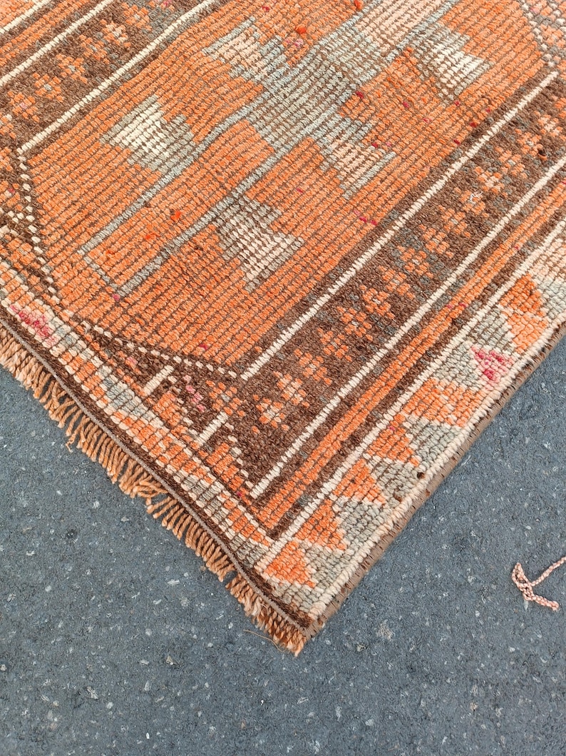 May include: A woven rug featuring a vibrant orange and brown geometric pattern with a central medallion design. The rug has a textured pile, a decorative border with repeating motifs, and short, light-colored fringe along the finished edge.