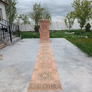 May include: A long, rectangular, vintage rug with a faded peach background and intricate geometric patterns in shades of brown and beige. The rug is displayed outdoors on a concrete surface, with a view of trees and a body of water in the background.