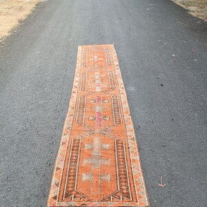 May include: A long, rectangular runner rug featuring a vibrant orange base color with intricate geometric patterns in shades of brown, cream, and pink. The rug has a repeating central motif and a fringed edge at the bottom, showcasing a woven texture.