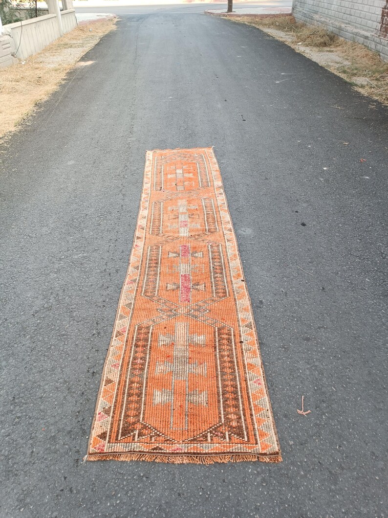 May include: A long, rectangular runner rug featuring a vibrant orange base color with intricate geometric patterns in shades of brown, cream, and pink. The rug has a repeating central motif and a fringed edge at the bottom, showcasing a woven texture.