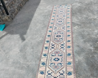May include: A long, rectangular rug with a central geometric pattern in shades of pink, blue, and beige. The rug is placed on a concrete surface, showcasing its intricate design and color palette. The rug's edges are bordered with a repeating pattern.
